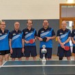 Acht Männer in blauer Sportkleidung stehen aufgereiht in einer Sporthalle. Sie halten Tischtennisschläger in der Hand.