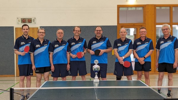Acht Männer in blauer Sportkleidung stehen aufgereiht in einer Sporthalle. Sie halten Tischtennisschläger in der Hand.