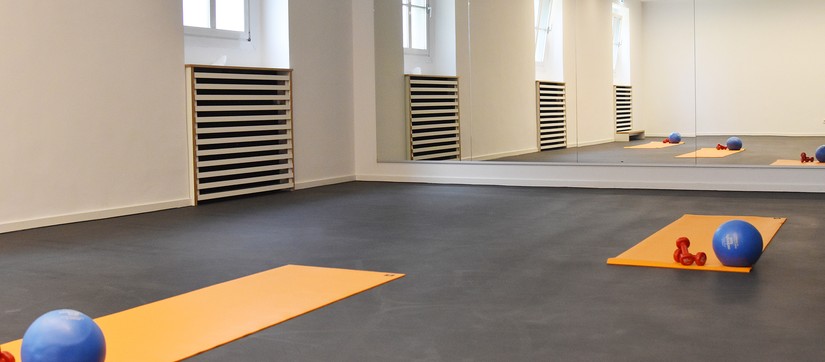 Sportraum mit schwarzem Boden, darauf liegen zwei orangene Matten und jeweils mehrere Sportutensilien